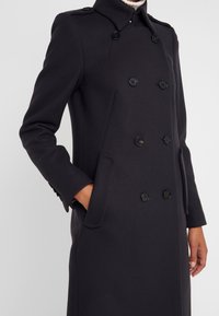 Abrigo azul marino de doble botonadura con cuello alto, que presenta botones negros, bolsillos laterales y un ajuste entallado. Tejido suave y cálido.