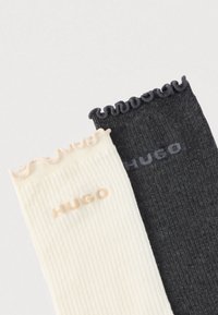 Calcetines acanalados en crema y gris oscuro; puños decorativos con volantes; logo "HUGO" en colores coordinados. Textura suave, diseño casual.