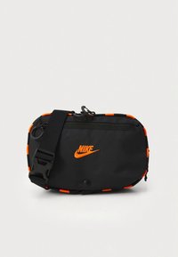 Bolsa de cintura preta da Nike com logótipo e acabamento em laranja, bolso frontal com fecho de correr, alça ajustável com fivela preta, sobre um fundo branco simples.