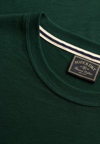T-shirt vert foncé à col rond avec une étiquette cousue indiquant « Superdry Premium Outfits New York 1954 » à l’intérieur du col.
