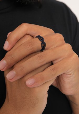 Navy blue chain-link ring made of flexible material, featuring interlocking shapes and a smooth texture, worn on a hand with natural nails.