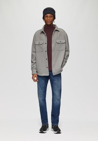 Grijze button-up jas met twee borstzakken over een bordeauxrode turtleneck, gecombineerd met blauwe jeans en zwarte sneakers, afgemaakt met een zwarte beanie.
