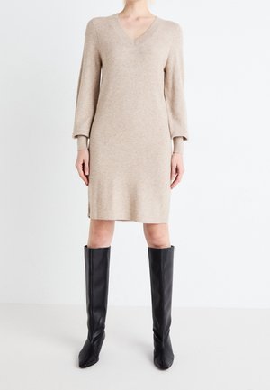 Beiges Strickkleid mit V-Ausschnitt und langen Ärmeln, gerade geschnitten mit seitlichen Schlitzen. Kombiniert mit knielangen schwarzen Stiefeln.