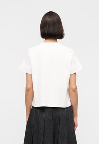 Γυναίκα με κοντά σκοτεινά μαλλιά που φοράει ένα απλό λευκό κοντομάνικο τοπ και μια μαύρη πλισέ φούστα, εμφανιζόμενη από πίσω πάνω σε λευκό φόντο.