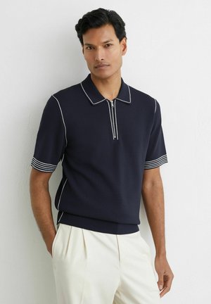 Férfi sötétkék rövid ujjú galléros pólóval, fehér szegéllyel és cipzárral, fehér pliszírozott nadrággal, fehér fal előtt állva.