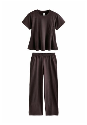 Junge Frau mit langen dunklen Haaren, die einen lockeren schwarzen Oberteil und schwarze Hose trägt, geht durch einen weißen Türrahmen in einem minimalistisch eingerichteten Raum mit Holzboden.