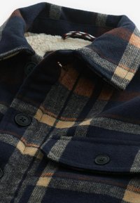 Veste en laine à carreaux bleu marine et marron avec col boutonné, boutons noirs et doublure polaire beige douce visible au niveau du col.