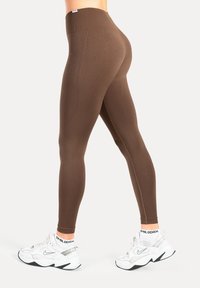 Leggings marroni a vita alta realizzati in materiale elastico, con una texture liscia e un design aderente. Scarpe da ginnastica bianche completano l'outfit.