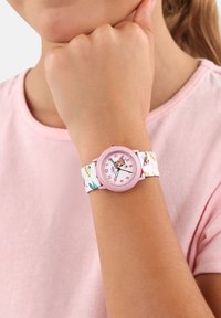 Roze horloge met een witte stoffen band met kleurrijke bloempatronen. De wijzerplaat heeft duidelijke cijfers en decoratieve accenten.