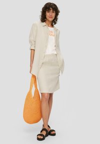 Beige Leinenhemd, weißes T-Shirt mit orangefarbenem Text, hellbeige Rock und schwarze Sandalen. Orange geflochtene Tasche mit großen Öffnungen.