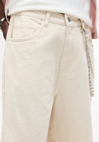 Pantaloni in denim color crema con superficie testurizzata, dotati di tasche frontali e dettaglio in catena argentata lucida sul lato.