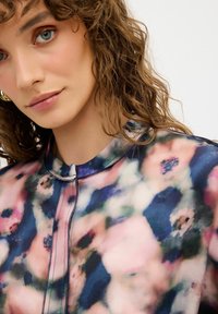 Blus med blommönster i mjukt, lätt material; har en rundad krage, framknäppning och en blandning av rosa, grön och blå nyanser.