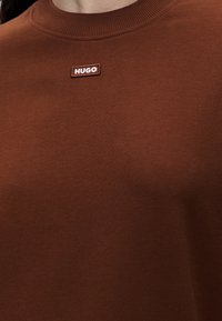 Gros plan d'un pull marron à col rond avec un petit patch rectangulaire étiqueté "HUGO" près du col.