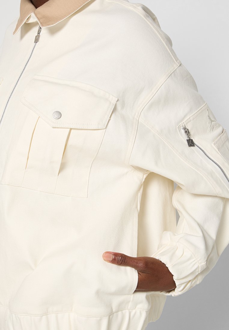 Chaqueta blanca texturada con cierre frontal de cremallera, dos bolsillos en el pecho y un cuello beige. Incluye puños elásticos y un bolsillo lateral con cremallera.