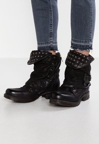 Botines de cuero negro con diseño holgado, cuello doblado con tachuelas, cremallera lateral y tacón de madera robusto. Detalle de superficie texturizada.