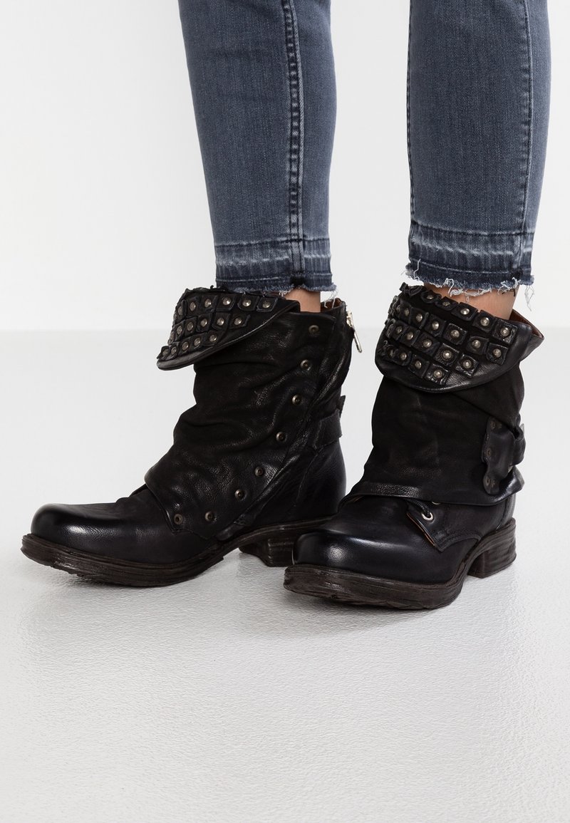 Botines de cuero negro con diseño holgado, cuello doblado con tachuelas, cremallera lateral y tacón de madera robusto. Detalle de superficie texturizada.