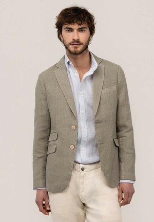 Hombre con blazer beige claro sobre una camisa de rayas azul claro con botones y pantalones blanco roto, de pie delante de un fondo simple.