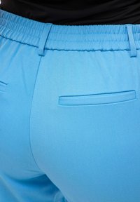 Lyseblå bukser med elastisk midjebånd. Har to baklommer og en jevn, teksturert stoff.