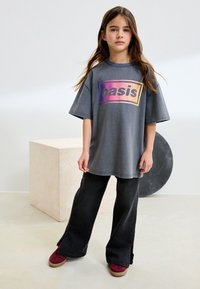 Niña joven con cabello largo y castaño que lleva una camiseta gris de gran tamaño con la palabra "oasis", vaqueros negros de pierna ancha y zapatillas rojas, parada en el interior con elementos geométricos como accesorios.