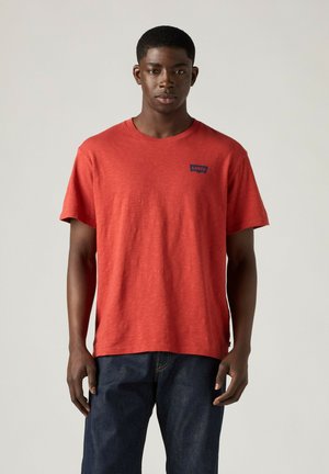 Joven que lleva una camiseta roja de Levi’s y vaqueros azul oscuro, de pie frente a un fondo liso y claro, mirando hacia adelante.