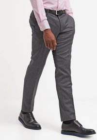 Graue, maßgeschneiderte Hose mit strukturiertem Stoff, schmaler Passform und Bügelfalte vorne. Kombiniert mit schwarzen eleganten Schuhen und einem schwarzen Gürtel.