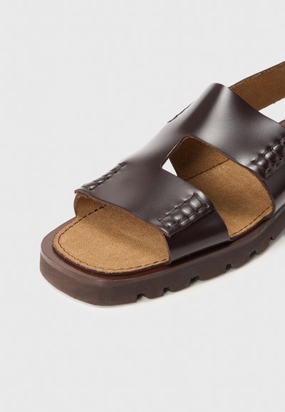 Sandalo marrone in pelle con due sezioni ritagliate e dettagli intrecciati. Presenta una soletta testurizzata e imbottita e una suola robusta e scura.