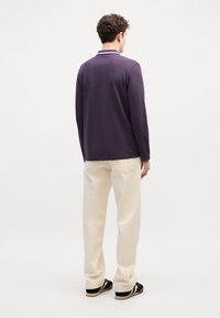 Πολυάριθμο πουκάμισο μακρυμάνικο σε σκούρο μωβ, με γιακά και λευκή ραφή; φοριέται με κρεμ παντελόνι φαρδύ και μαύρες παντόφλες.