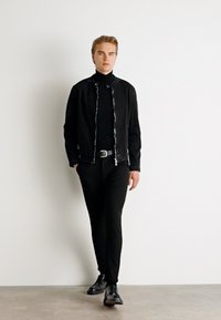 Black zip-up jacket with a textured finish, layered over a black turtleneck, paired with black pants and sleek black shoes.