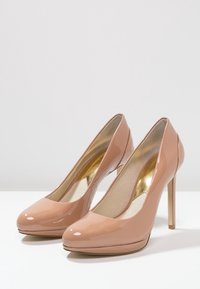 Talons hauts en cuir verni nude avec un bout rond, un talon aiguille mince et une semelle intérieure rembourrée. Accents intérieurs dorés visibles.