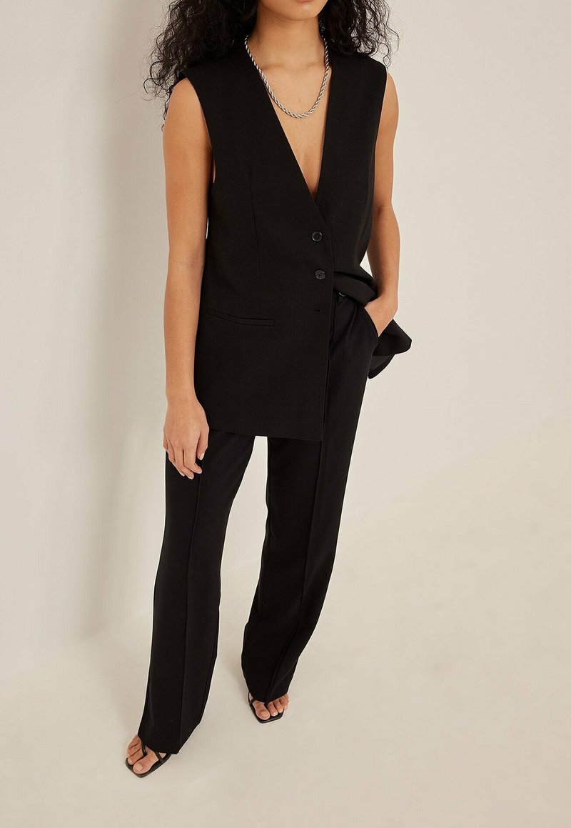 Čierny bezrukávový blejzer s hlbokým výstrihom do V, zapínaním na jeden gombík a prednými vreckami, skombinovaný s čiernymi priliehavými nohavicami a sandálmi.