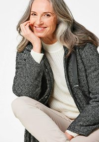 Zwart-wit Visgraat textuurjas met drukknoopsluitingen en zwarte accenten, gedragen over een crèmekleurige kabelgebreide trui en lichtbeige broek.