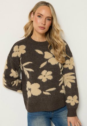 Vrouw met lang blond haar, die een donkerbruine trui draagt met grote beige bloemenpatronen en blauwe spijkerbroek, staand voor een effen achtergrond.