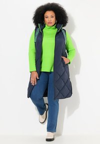 Granatowy długi kamizelka ze wzorem rombowym, z zielonymi paskami na tekst, noszona na jasną zieloną golf, w połączeniu z niebieskimi dżinsami i kremowymi butami.