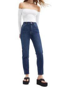 Haut blanc dénudé des épaules, jean taille haute bleu foncé, et sandales à plateforme noires avec des bretelles épaisses et une semelle texturée.