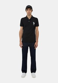 Polo nero con maniche corte e due bottoni, con un logo di un cavallo blu chiaro e il testo "USPA". Abbinato a jeans scuri e scarpe bianche.
