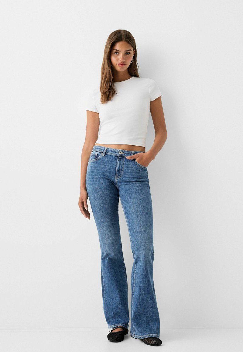 Giovane donna con lunghi capelli castani, che indossa un crop top bianco a maniche corte, jeans a vita alta svasati blu e scarpe nere, in piedi.
