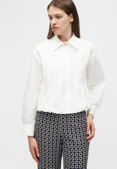 Femme portant un chemisier blanc à manches longues avec un col large et un pantalon noir à motifs avec des dessins circulaires blancs et orange.