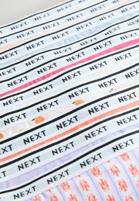 Gros plan sur des bandes de tissu avec le texte noir "NEXT" répété, ainsi que des rayures de différentes couleurs et de petits motifs orange et rouges.