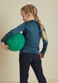 Långärmad teal atletisk tröja i kombination med mörkblå byxor, håller en grön basketboll med svarta ränder, sedd bakifrån.