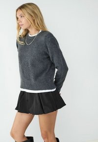 Grauer Wollmischpullover, Rundhalsausschnitt, über einem weißen Oberteil getragen, kombiniert mit einem schwarzen gefältelten Minirock und knielangen schwarzen Stiefeln.