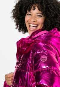 Chaqueta acolchada fucsia brillante con cierre de cremallera en el frente, capucha adjunta y una insignia en el hombro izquierdo. El material tiene una textura suave y reflectante.