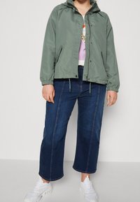 Light green, water-resistant jacket with a hood, elastic cuffs, and drawstring hem, paired with dark blue wide-leg jeans and white sneakers.
