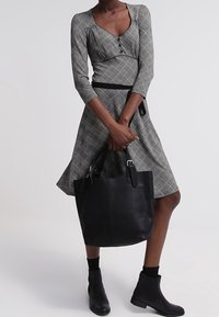 Svart läder tote-väska med dubbelhandtag, slät yta och silverdetaljer. Modellen bär en grå rutiga klänning med trekvartslånga ärmar.