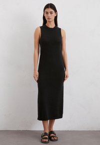 Schwarzes, gestricktes, ärmelloses Kleid mit hohem Ausschnitt, schlankem Schnitt und knielanger Länge. Getragen mit schwarzen Sandalen mit goldenen Akzenten.