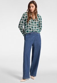 Blusa verde a fantasia con un design a catena bianca; pantaloni in denim larghi con dettagli a bottoni sui lati; tacchi beige.