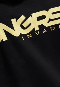 Svart tygskjorta med djärv, guldtext som står "NGRS INVADED." Designen har en slät, platt yta med ett modernt typsnitt.