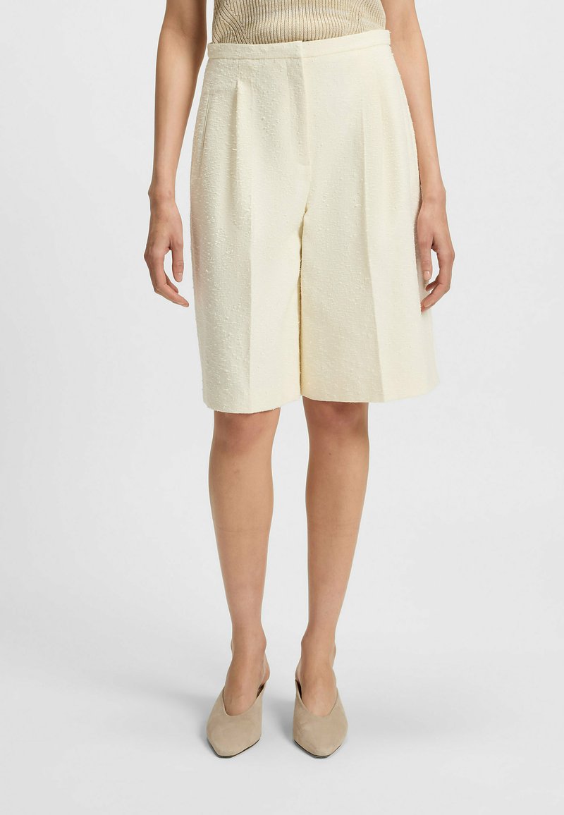 Kermanväriset räätälöidyt shortsit, joissa on aikaa kestävää kangasta, ominaisuuksina laskokset ja suora leikkaus reiden puoliväliin asti, yhdistettynä vaaleanbeigeihin kenkiin.