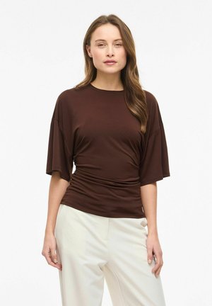 Frau mit langen braunen Haaren, die ein braunes, kurzärmeliges, gerafftes Oberteil und weiße, hoch taillierte Hosen vor einem weißen Hintergrund trägt.