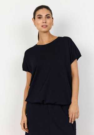 Kobieta o ciemnych włosach związanych do tyłu, ubrana w luźną, czarną bluzkę z krótkim rękawem i dopasowaną czarną spódnicę na jednolitym białym tle.