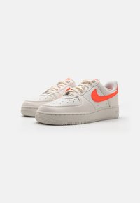 Zapatillas Nike Air Force 1 en beige con acentos en naranja brillante. Presentan una caja de dedos perforada, suela de goma beige y un diseño con cordones.